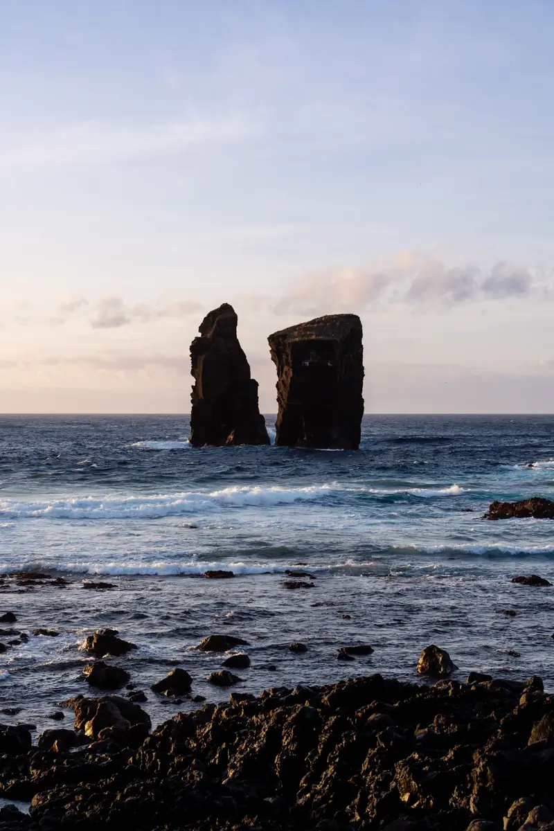 Two large sea stacks rising from the ocean off a rocky coastline, with waves breaking around them at sunset.
