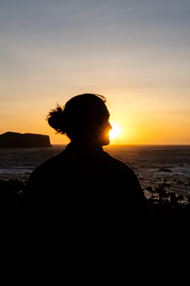Silhouette of a person looking out at the ocean during sunset, with the sun low on the horizon.