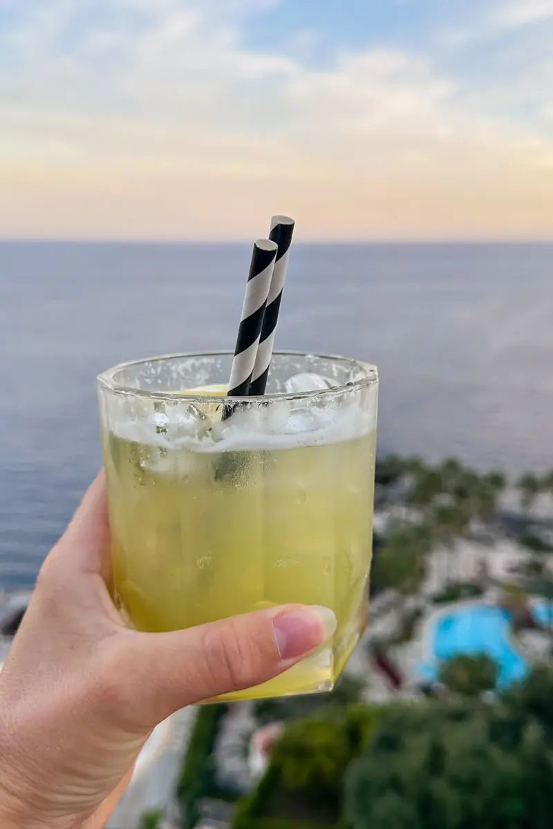 A hand holds a glass of yellow cocktail with two striped paper straws against a backdrop of the ocean at sunset. A resort pool and palm trees are visible below from an elevated viewpoint.