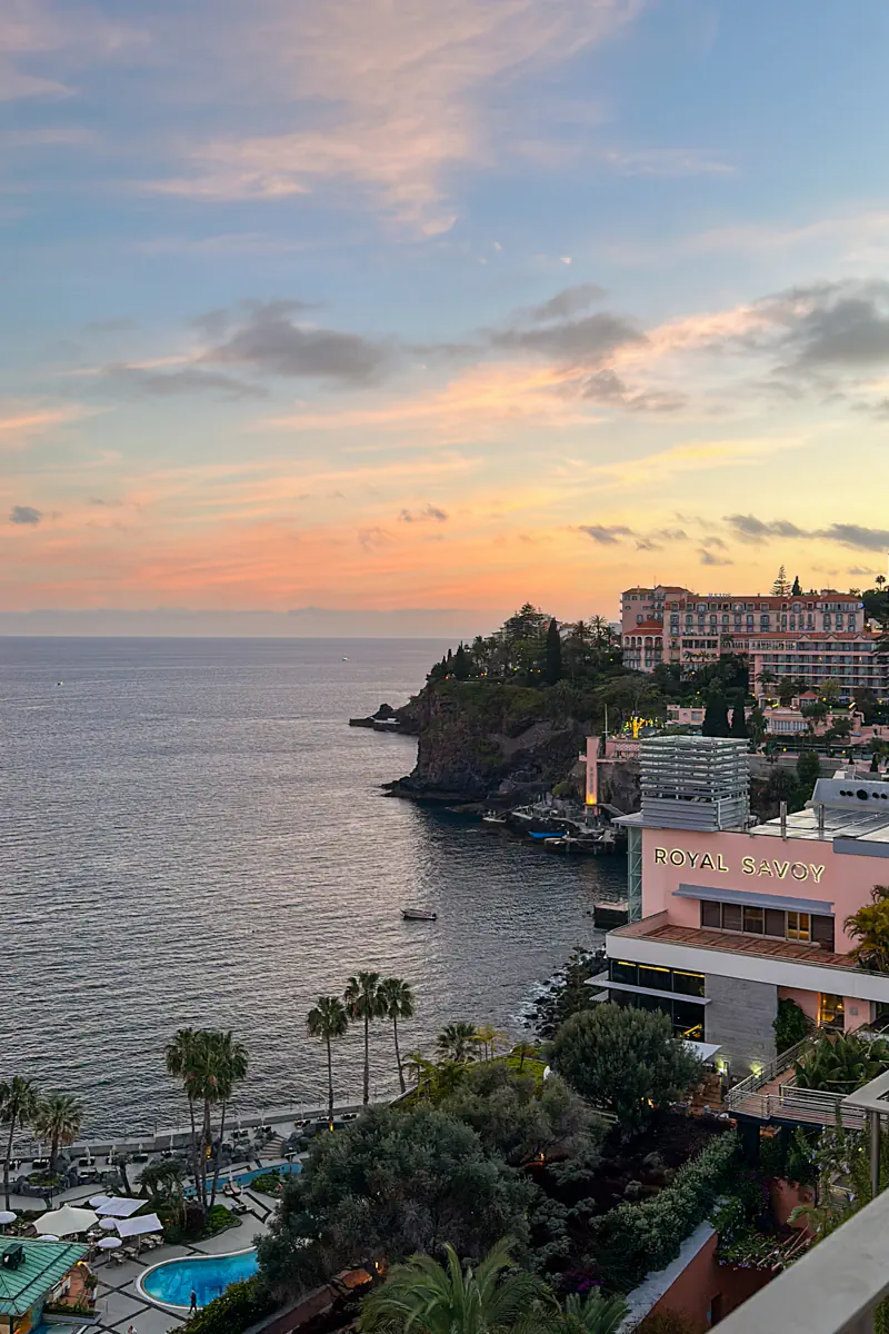 A coastal hotel complex sits on a rocky cliff overlooking the ocean at sunset, with soft pink and orange clouds in the sky. Palm trees and a pool terrace are visible below, and the building displays the name “Royal Savoy.”