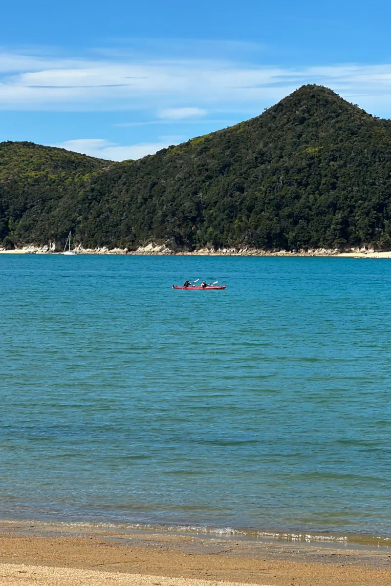 Two people kayak across calm blue water near a sandy beach with forested hills rising in the background. The peaceful bay and gentle waves highlight a relaxed outdoor adventure setting.