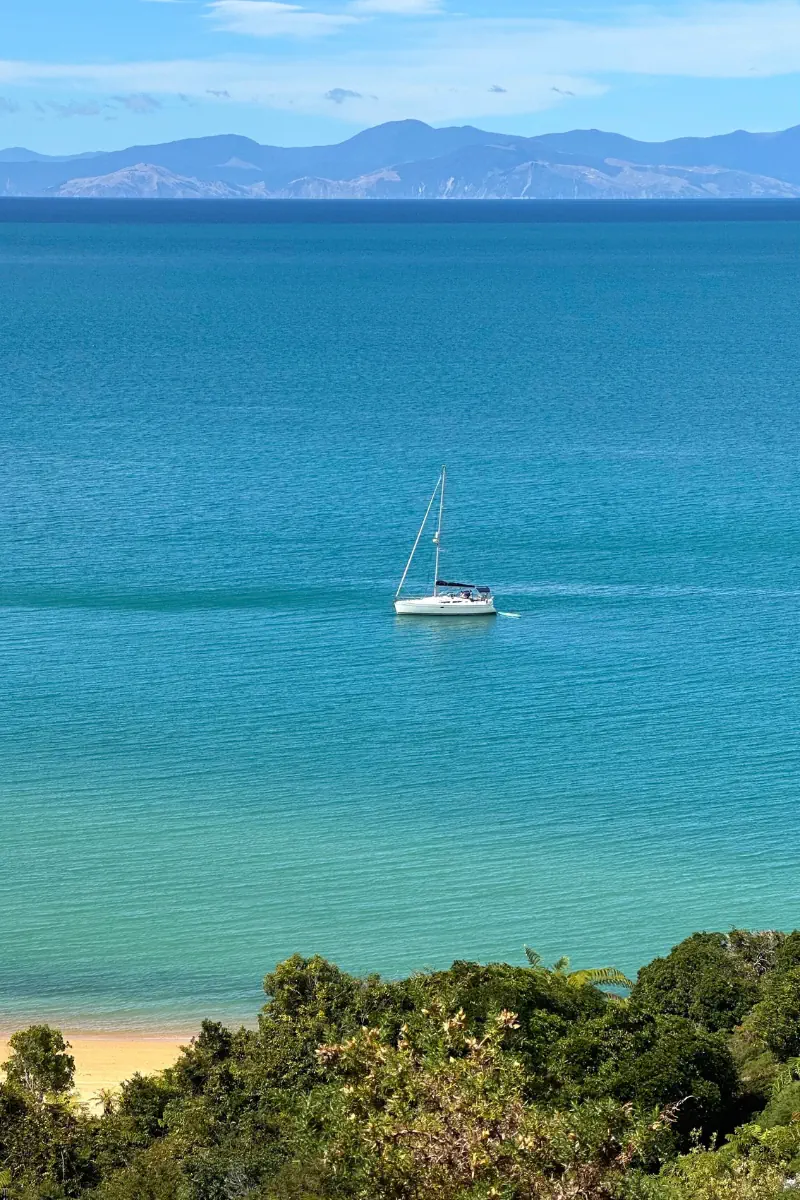A lone sailboat drifts across calm turquoise water with distant mountains lining the horizon under a soft blue sky. Lush green foliage and a sandy shoreline frame the peaceful coastal view from above.