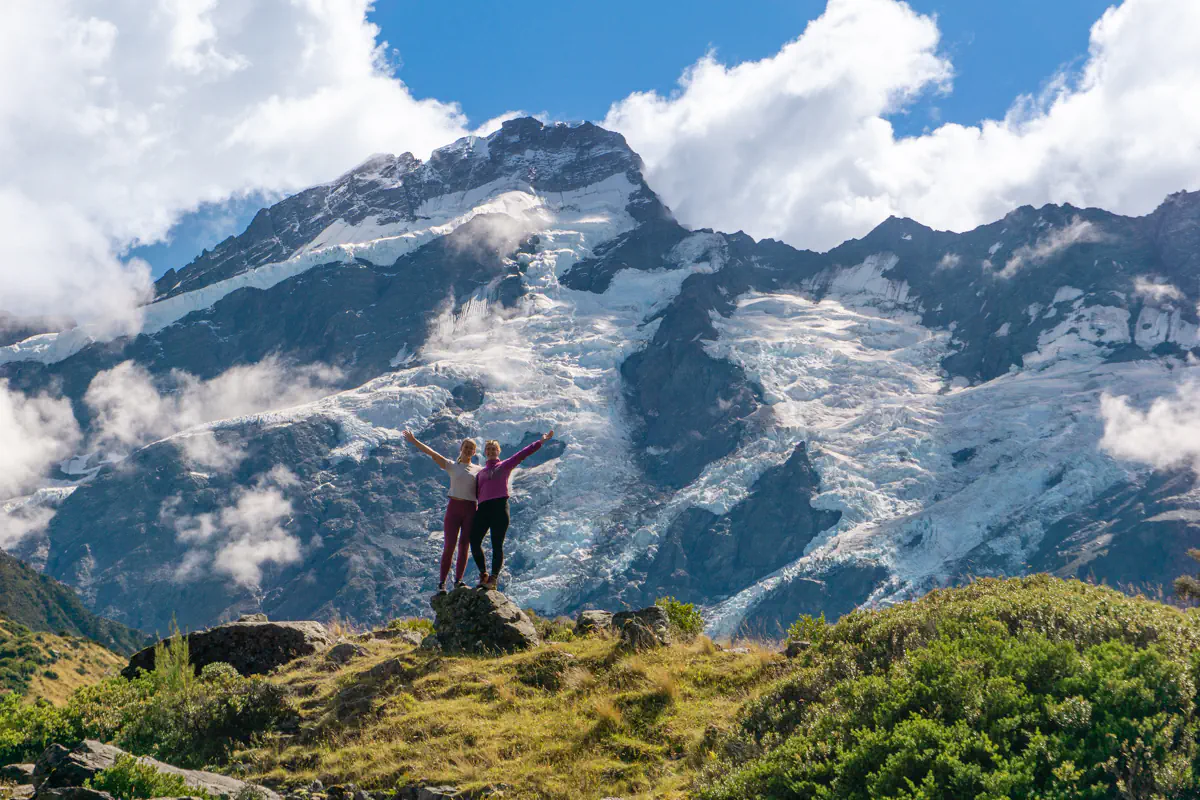Two travellers stand on a rocky outcrop with arms raised in front of a towering snow capped mountain and glacier surrounded by drifting clouds. The dramatic alpine landscape highlights the scale of the peaks and the sense of adventure in the moment.