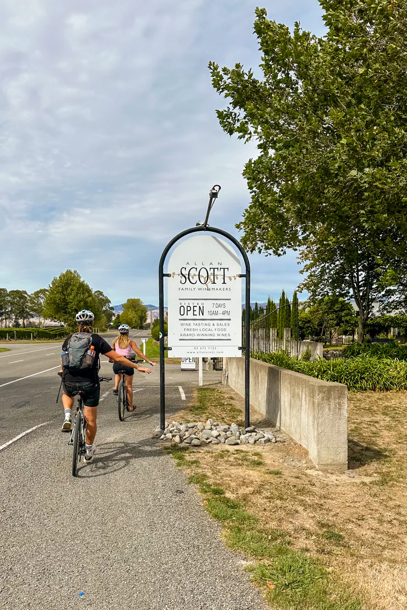Two cyclists ride past a roadside sign that reads Allan Scott Family Winemakers. The scene shows a scenic bike ride through wine country with vineyards and trees lining the road.