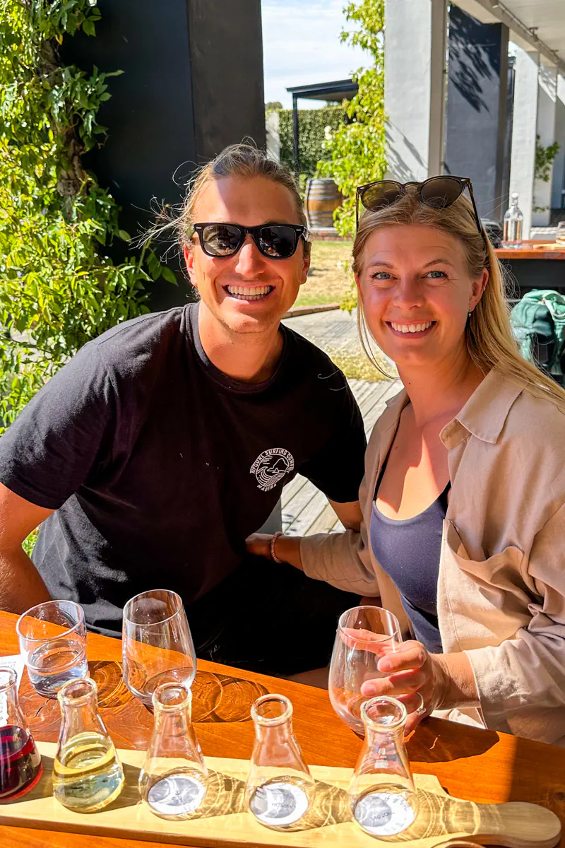 A couple sits at an outdoor table smiling with wine glasses and a tasting flight of small carafes in front of them. The image captures a winery visit and enjoying wine together in a sunny setting.