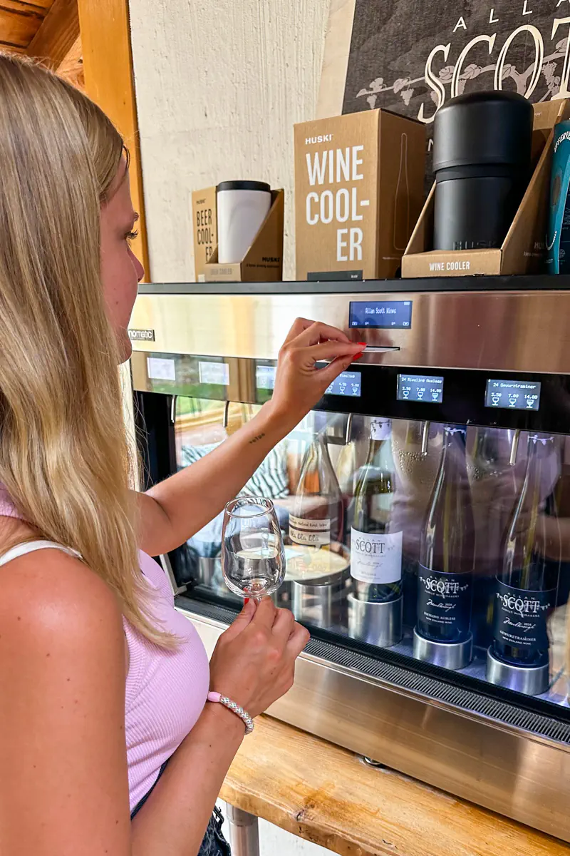 A woman pours a glass of wine from a self serve tasting machine with bottles labeled behind glass and boxes reading wine cooler on display.