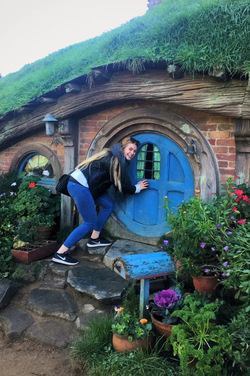 A woman steps up to a round blue door on a hobbit style house with a grass covered roof and colourful flowers around the entrance. The small curved doorway and rustic details create a whimsical garden setting.
