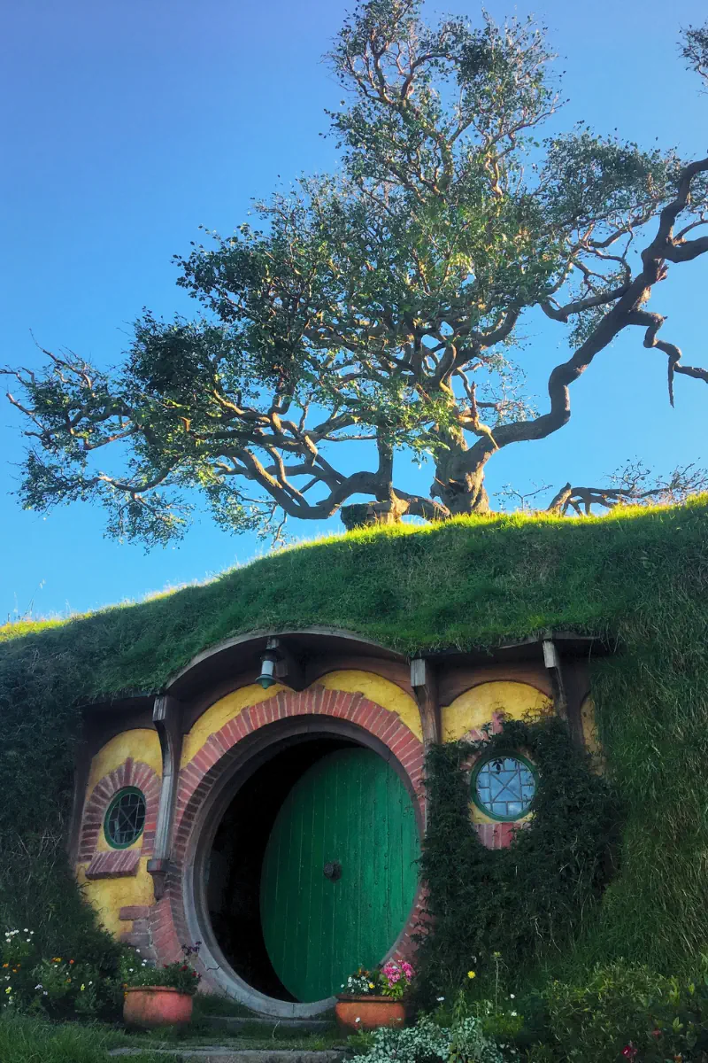 A hobbit house with a green round door and yellow walls sits beneath a grassy hill topped by a large tree. The curved architecture and lush surroundings give it a storybook appearance.