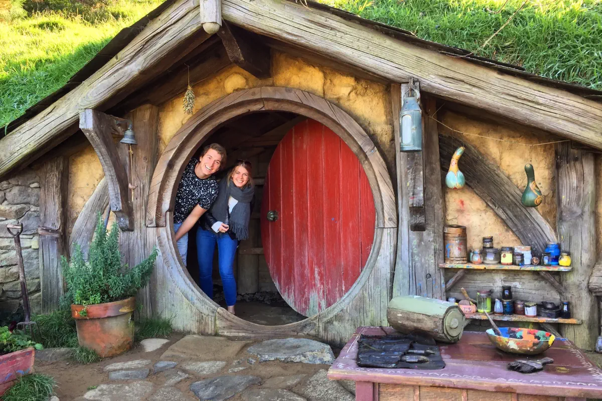 Two people smile while standing in the round doorway of a hobbit house with a red door and rustic wooden beams. Shelves with jars and tools sit beside the entrance, adding to the cozy lived in feel.