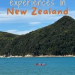 Two people kayak across calm turquoise water with forested hills in the background under a clear sky. Text reads 11 bucket list experiences in New Zealand.