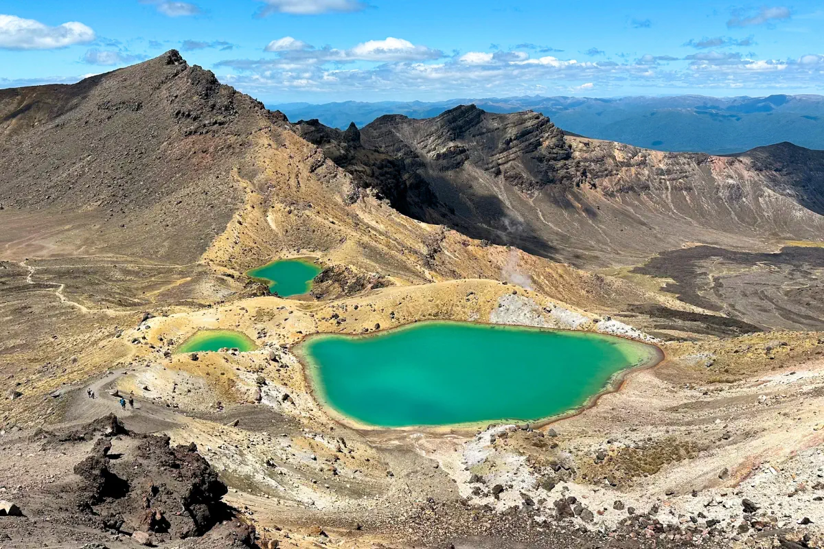Turquoise alpine lakes sit in a volcanic crater surrounded by rugged brown mountains and winding hiking trails. The bright blue water contrasts sharply with the barren rocky landscape under a partly cloudy sky.