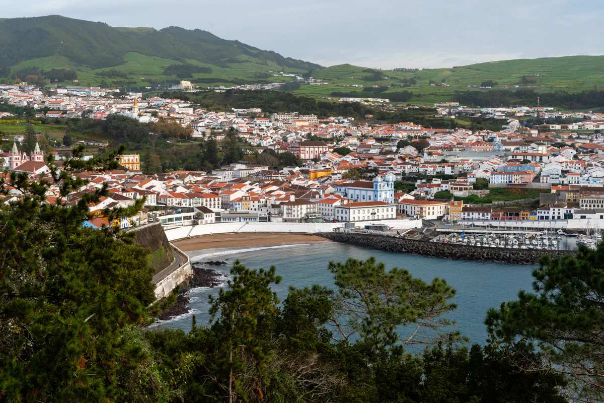Coastal view of Angra do Heroísmo with red rooftops, a marina, a beach, historic churches, and green hills surrounding the town on Terceira Island.