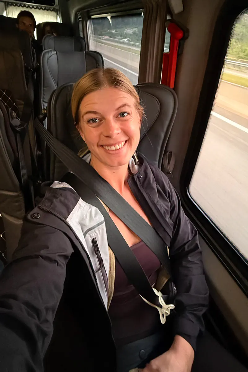 Woman smiling from a seat inside a shuttle bus with a seatbelt fastened while traveling along a highway.