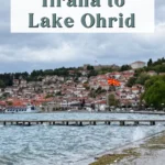Cloudy lakeside promenade in Ohrid with waves crashing along the shore, hillside homes, and a North Macedonian flag above the waterfront. Text overlay reads "HOW TO GET FROM Tirana to Lake Ohrid."