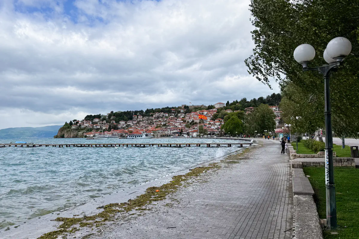 Lakeside promenade in Ohrid with waves crashing along the shore and a hillside town covered in red-roofed buildings under cloudy skies. A large North Macedonian flag waves above the waterfront near the centre of town.
