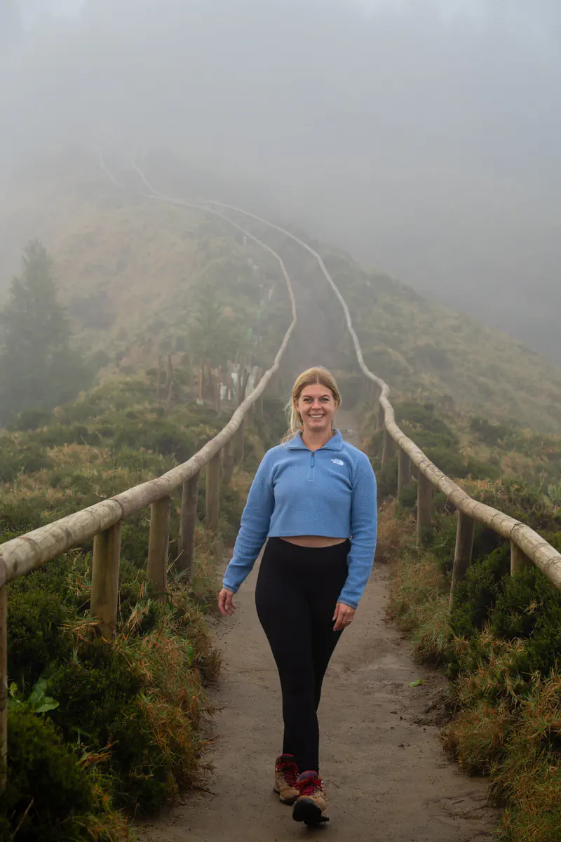 A smiling woman walks toward the camera on a misty ridge trail surrounded by low vegetation and wooden railings.