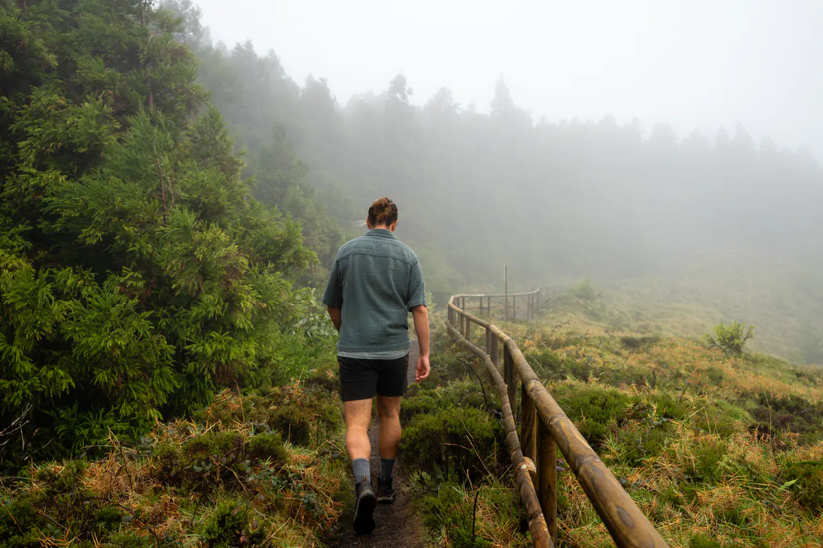 A man walks along a narrow dirt trail bordered by wooden railings with dense greenery on one side and thick fog ahead softening the landscape.