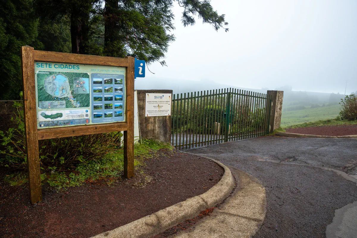 A wooden information board labeled Sete Cidades stands beside a metal gate overlooking a foggy green landscape beyond.