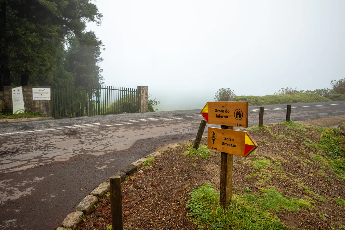 Trail signs point toward Grota do Inferno at 1.3 km and Serra Devassa at 2.5 km beside a quiet road surrounded by mist and low vegetation.