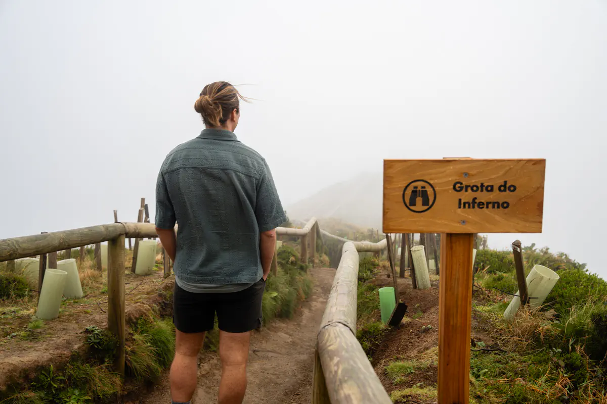 A man stands on a foggy viewpoint beside a wooden sign reading Grota do Inferno looking out over a mist covered landscape.