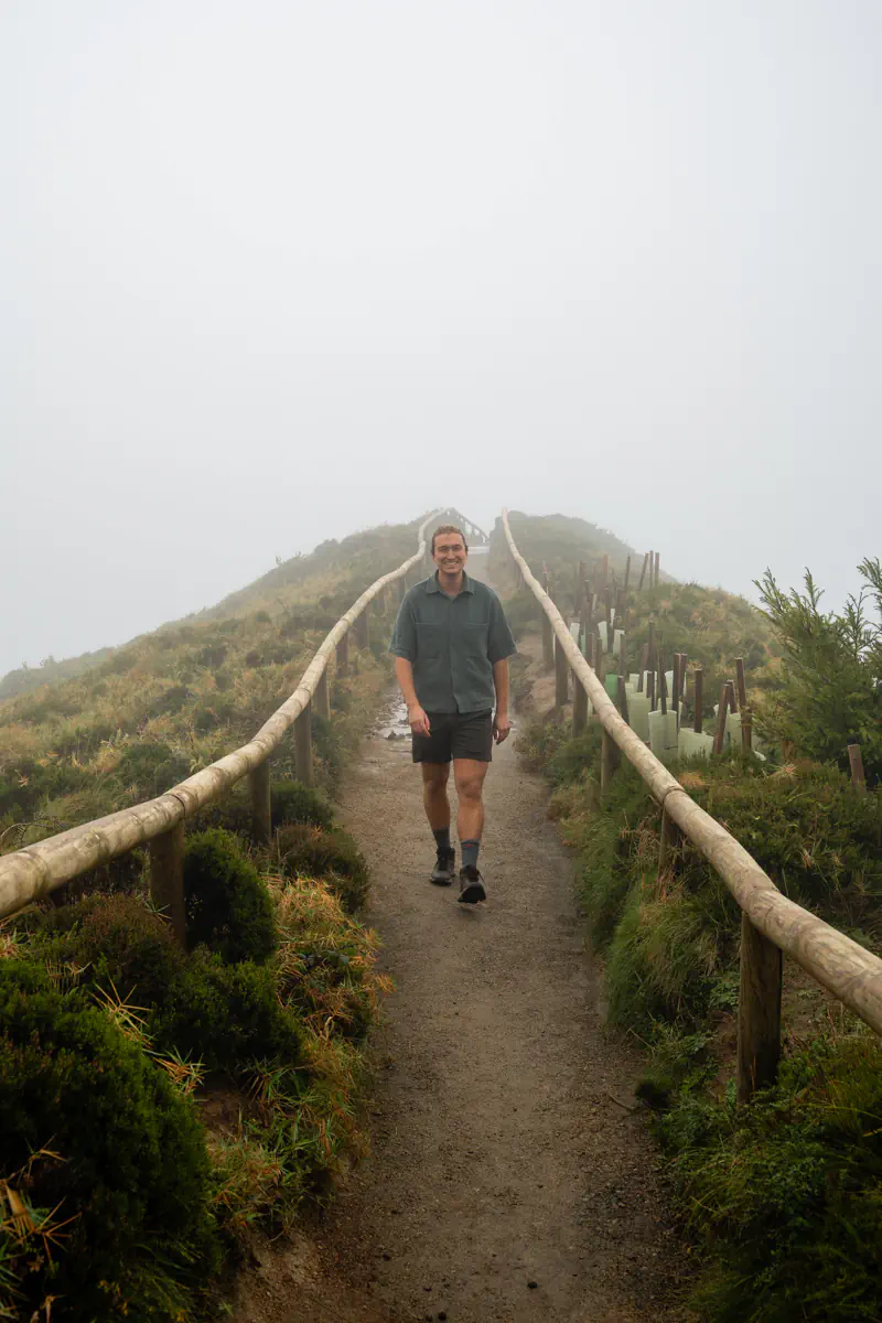 A smiling man walks toward the camera along a foggy ridge trail bordered by wooden railings and small planted trees.