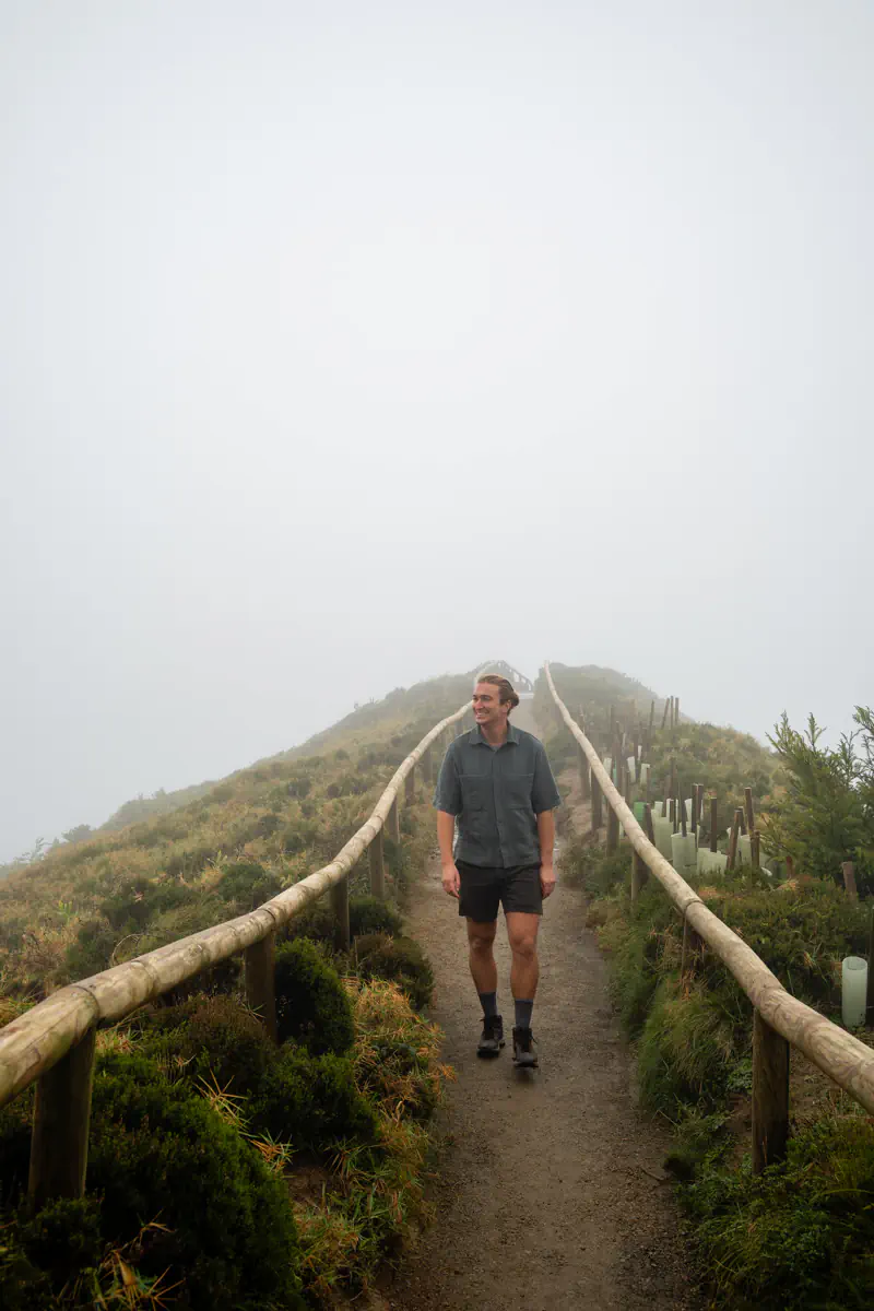 A man walks along a narrow ridge trail bordered by wooden railings with low green shrubs fading into thick fog around him.