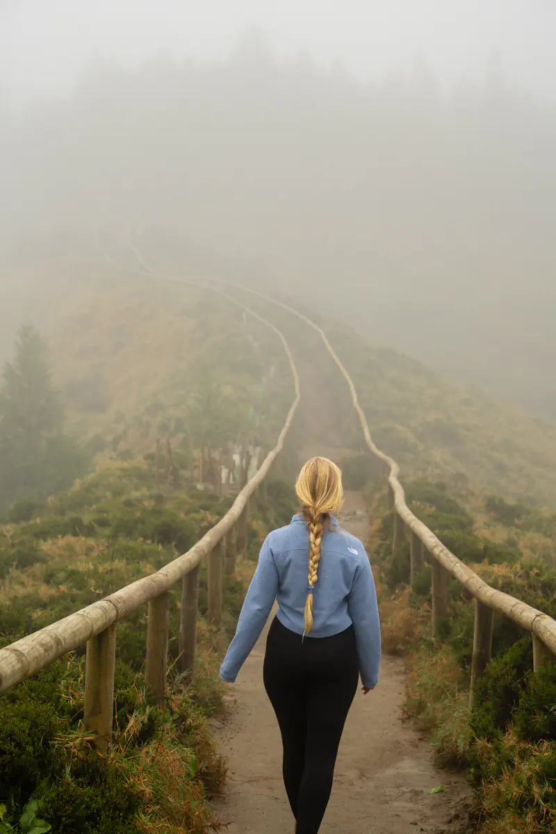 A woman walks down a narrow ridge trail with wooden railings as the winding path fades into heavy fog ahead.