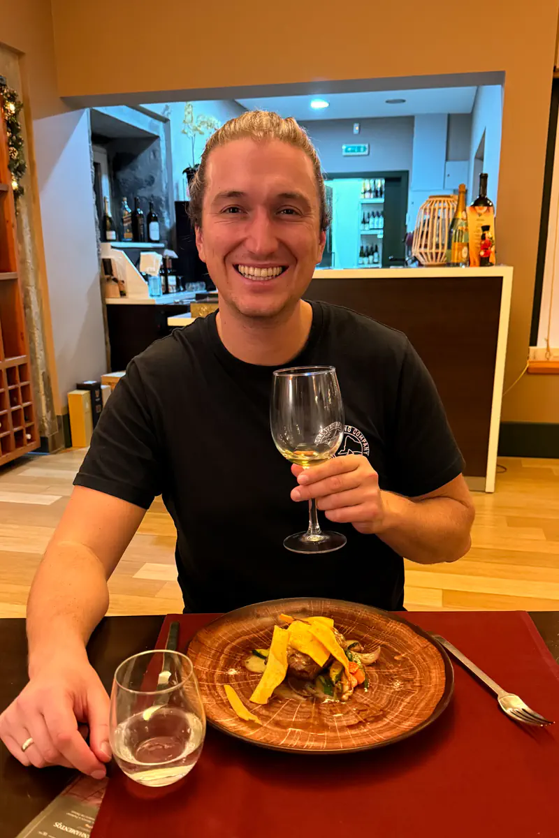 Man smiling while holding a glass of white wine at a restaurant table with a plated dinner in front of him.