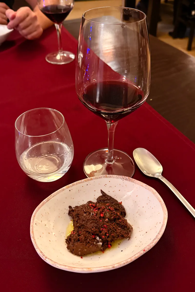 Dark chocolate dessert topped with pink salt flakes served in a white bowl beside a glass of red wine on a restaurant table.