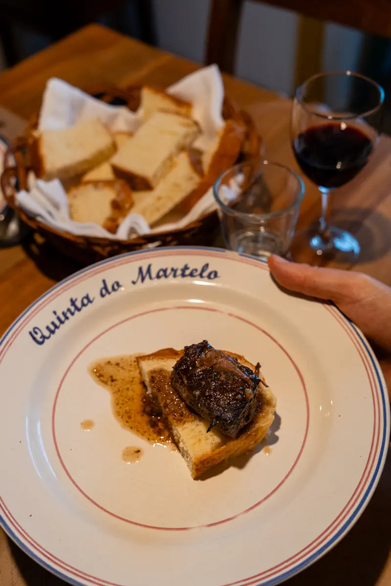 Small piece of braised meat served on a slice of bread with sauce on a white plate labelled “Quinta do Martelo,” beside a basket of bread and a glass of red wine.