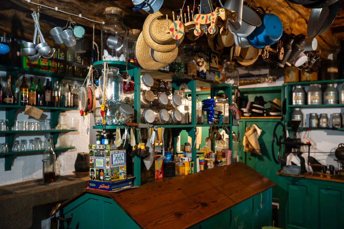 Shelves filled with vintage kitchen tools, glass jars, bottles, hats, and hanging cookware inside a traditional Azorean kitchen museum with teal wooden cabinets and a wooden counter.