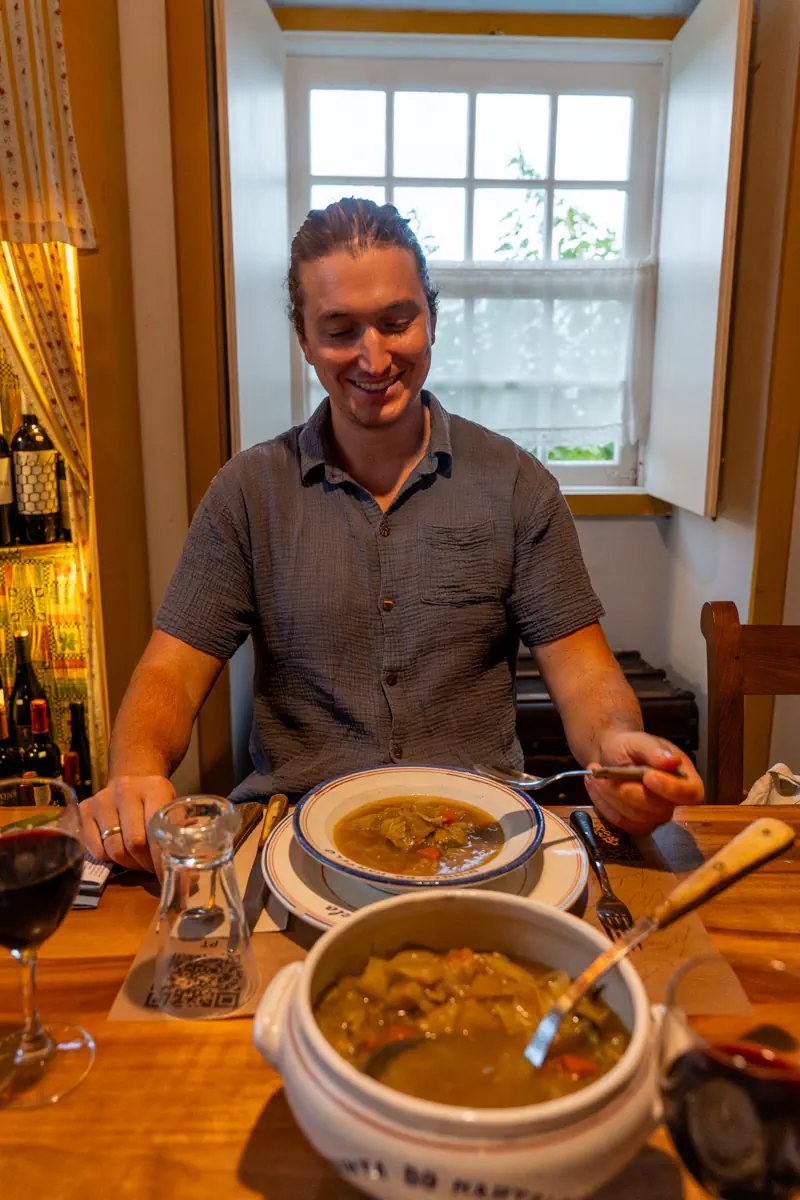 Man smiling while seated at a wooden dining table with a bowl of traditional Azorean soup and a glass of red wine in a cosy restaurant.