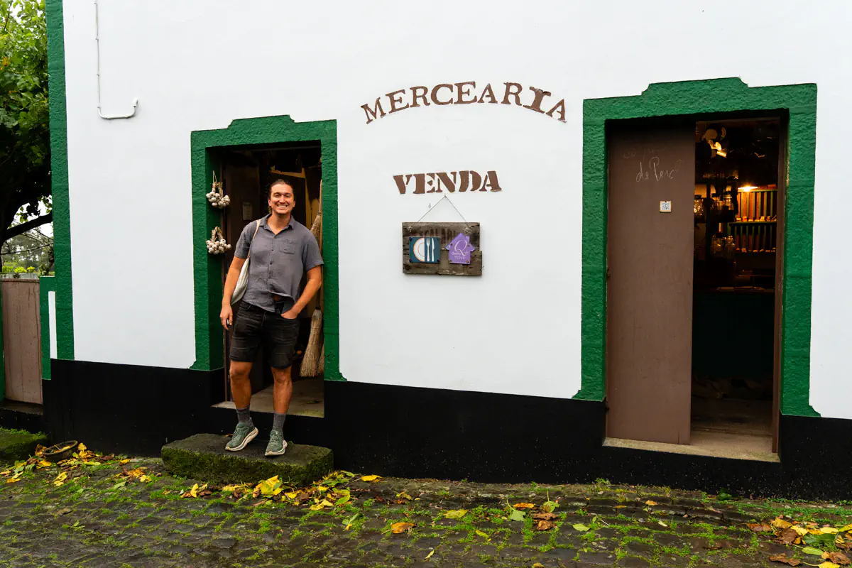 Man standing in the doorway of a white building with green trim and signs reading “Mercearia” and “Venda” on a rainy cobblestone street in the Azores.