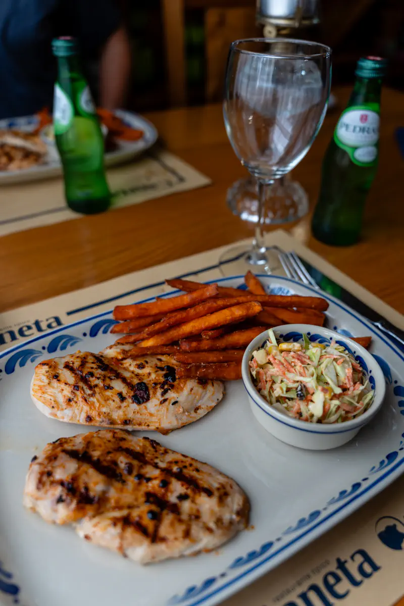 Grilled chicken breasts served with sweet potato fries and coleslaw on a white restaurant plate beside sparkling water bottles and wine glasses.