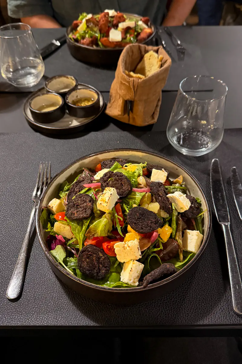 Large salad topped with blood sausage slices, cheese, tomatoes, oranges, and pickled onions served on a restaurant table with bread and dipping sauces.