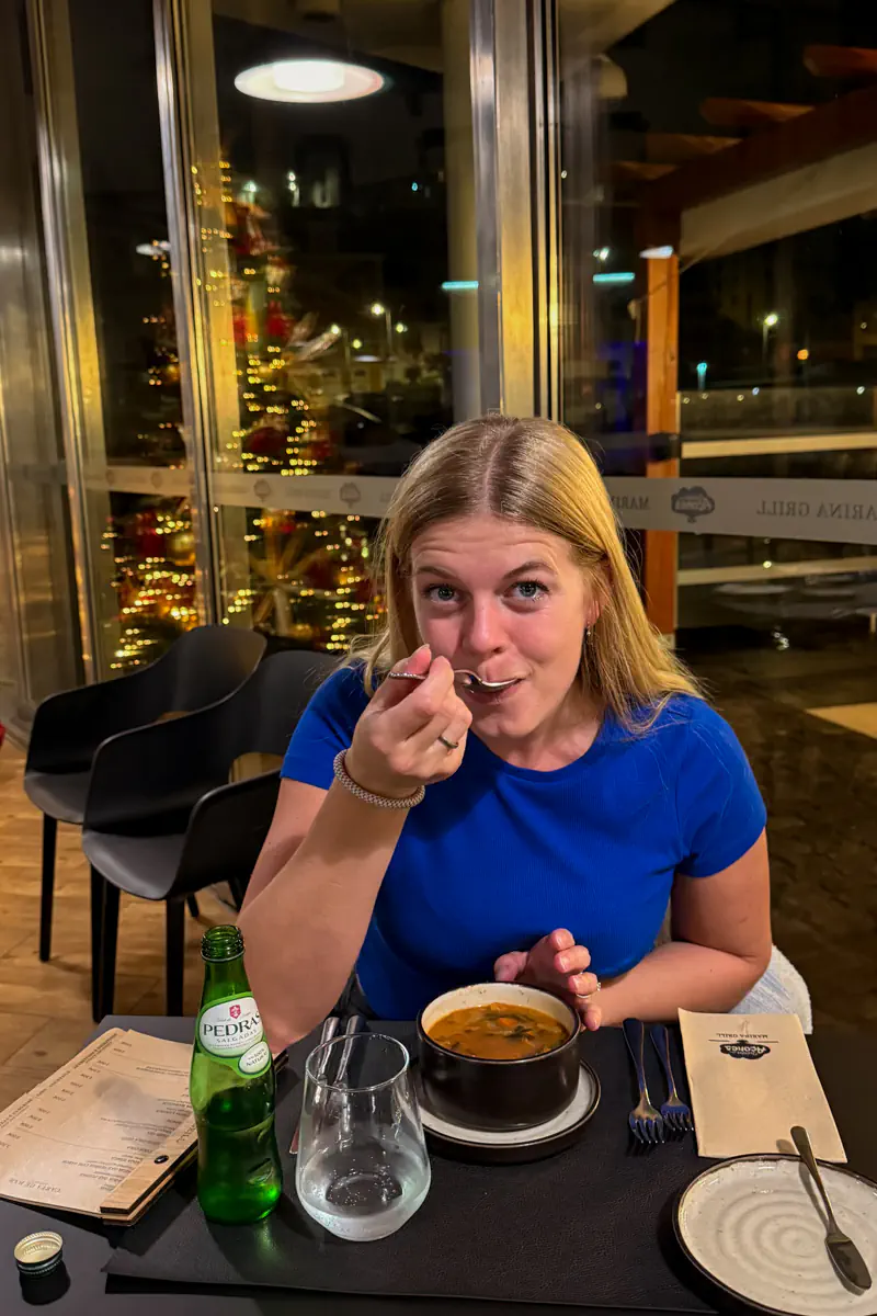 Woman eating soup at a restaurant table beside a bottle of sparkling water, with Christmas lights reflected in the window behind her.