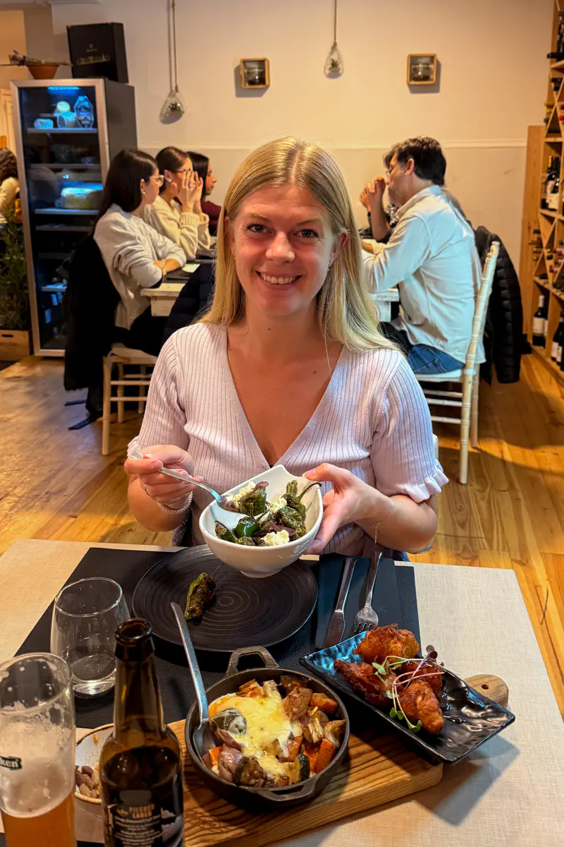 Woman smiling while holding a bowl of grilled spicy peppers at a restaurant table filled with tapas dishes and drinks.