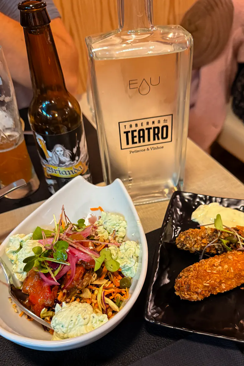 Tapas dishes including crispy croquettes, salad with pickled onions and herbs, and drinks on a restaurant table beside a bottle labelled “Taberna do Teatro.”