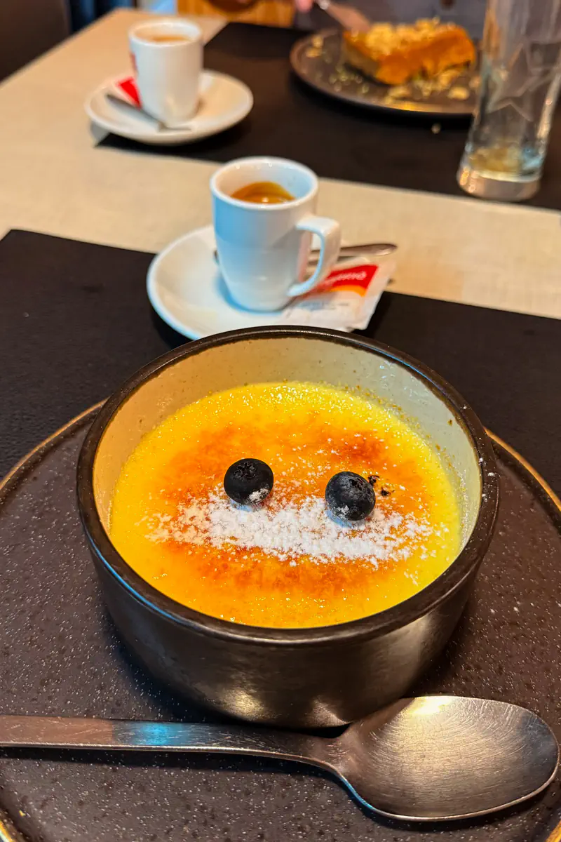 Crème brûlée topped with powdered sugar and blueberries served in a dark bowl beside espresso cups on a restaurant table.