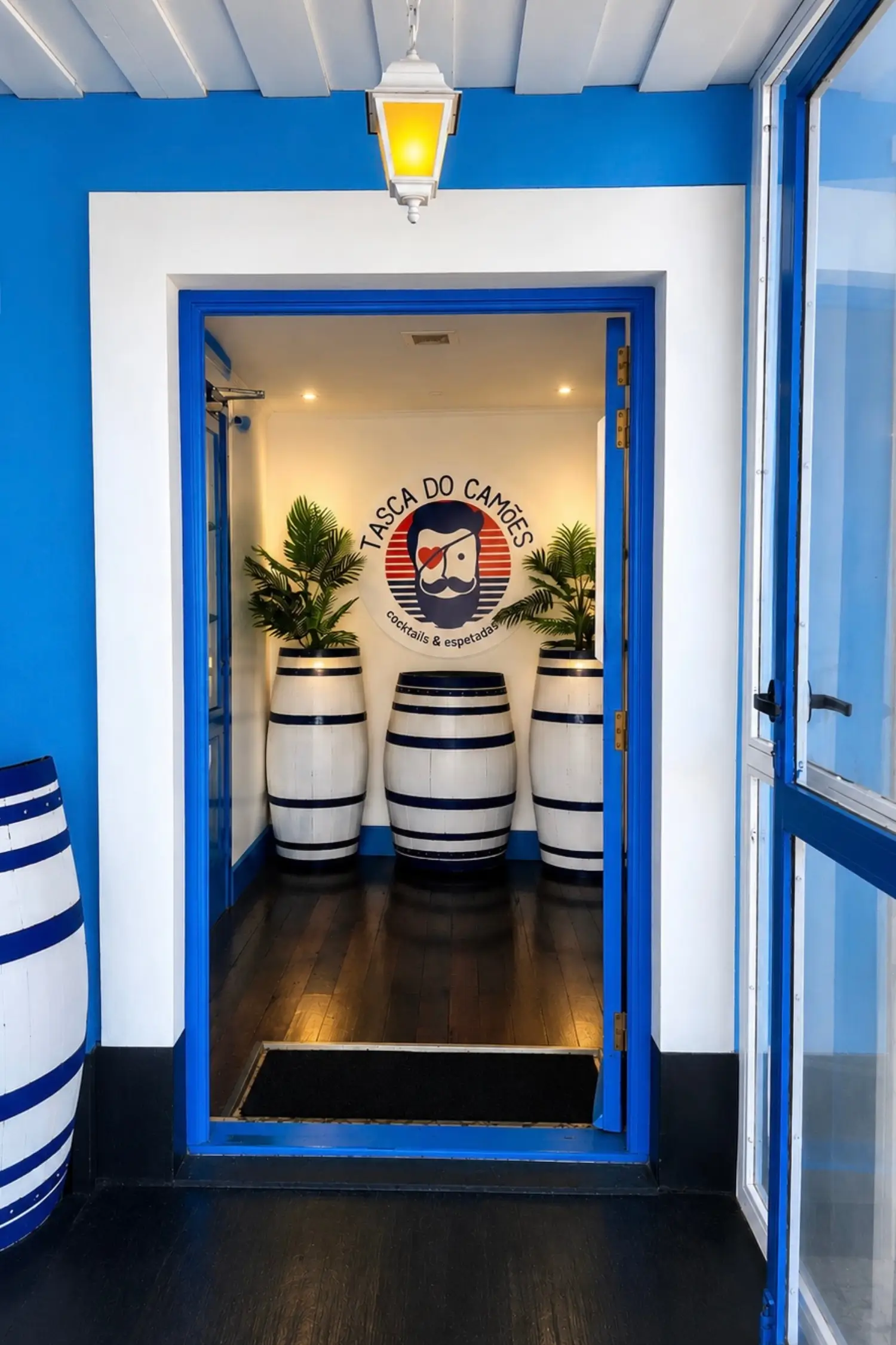 Bright blue and white entrance to Tasca do Camões restaurant with decorative barrels, tropical plants, and a wall logo reading "TASCA DO CAMÕES cocktails & espetadas."