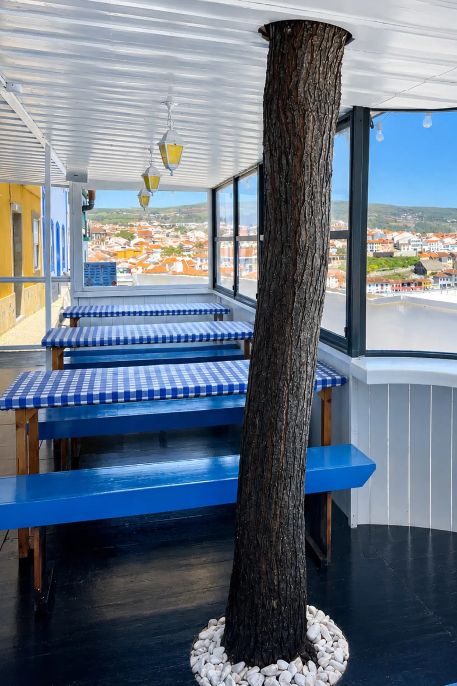 Covered restaurant seating area with blue checkered picnic tables and benches overlooking a town with red-roofed buildings. A tree trunk rises through the centre of the restaurant surrounded by white walls.