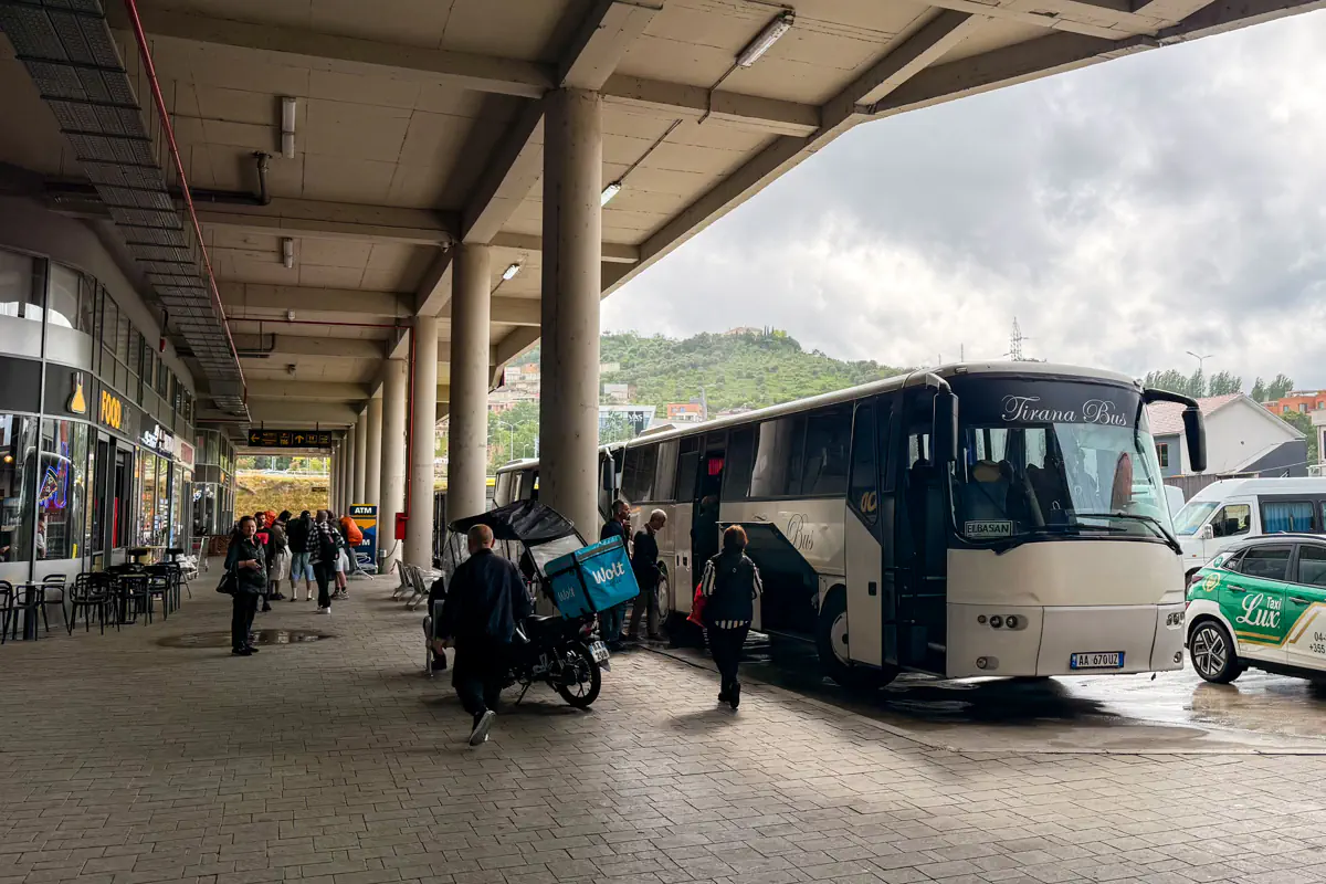 White Tirana Bus coach parked at a covered bus station with travellers boarding and walking through the terminal on a cloudy day.