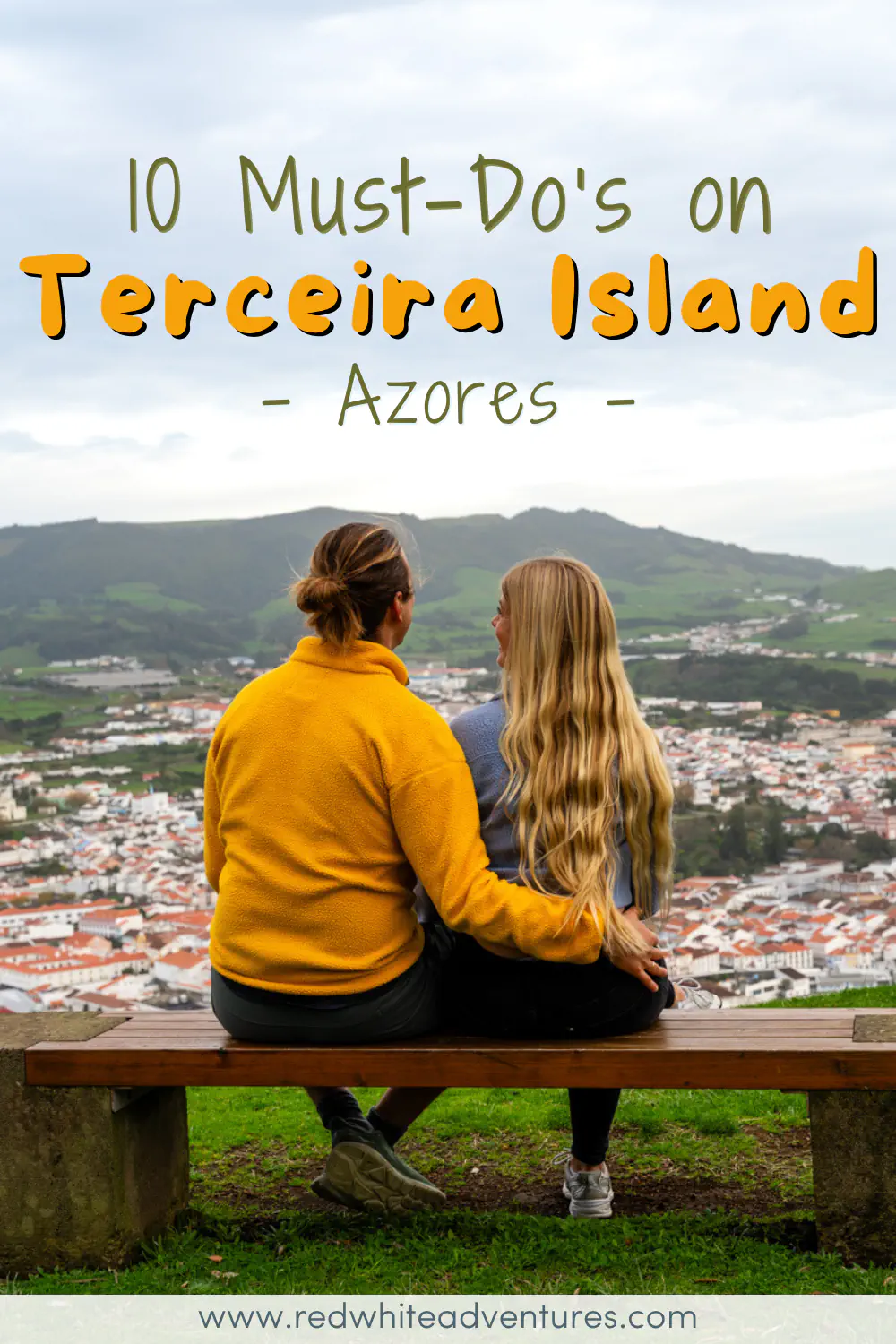 A couple sits side by side on a wooden bench at a hilltop viewpoint on Terceira Island, one with an arm around the other, looking out over the white buildings and orange rooftops of Angra do Heroísmo with green hills rising behind the city. Text overlay reads "10 Must-Do's on Terceira Island, Azores."