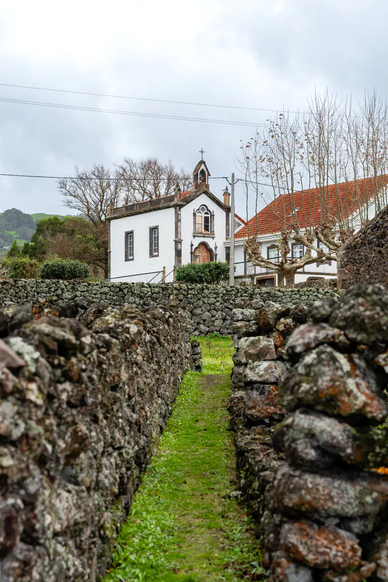 A narrow grass path runs between two lava stone walls at a winery on Terceira Island, leading toward a small white chapel with a cross and ornate wooden doorway, framed by bare trees under a cloudy sky.