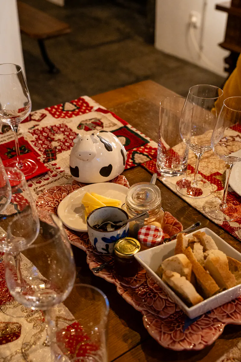 A rustic wooden table set for a cheese tasting at a winery on Terceira Island, with slices of yellow cheese, jam, bread sticks, small condiment jars, wine glasses, and a ceramic cow figurine on a decorative floral tray.