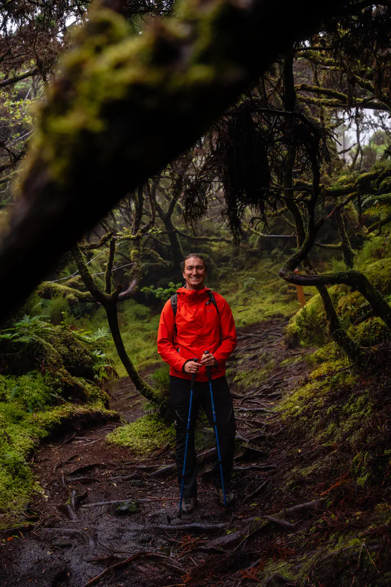 A smiling hiker in a dark orange rain jacket stands on a root-covered trail deep in a misty moss-draped forest on Terceira Island, holding trekking poles while twisted trees and vivid green ferns surround them on all sides.