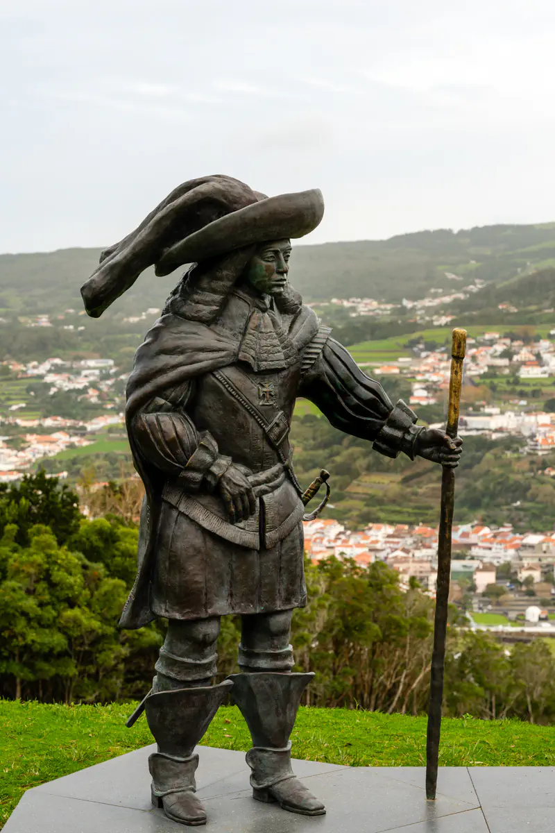 A bronze statue of a historical figure in a wide-brimmed hat, cloak, and sword stands on a hilltop viewpoint above Angra do Heroísmo on Terceira Island, with the town's terracotta rooftops and green volcanic hills spreading out behind it.