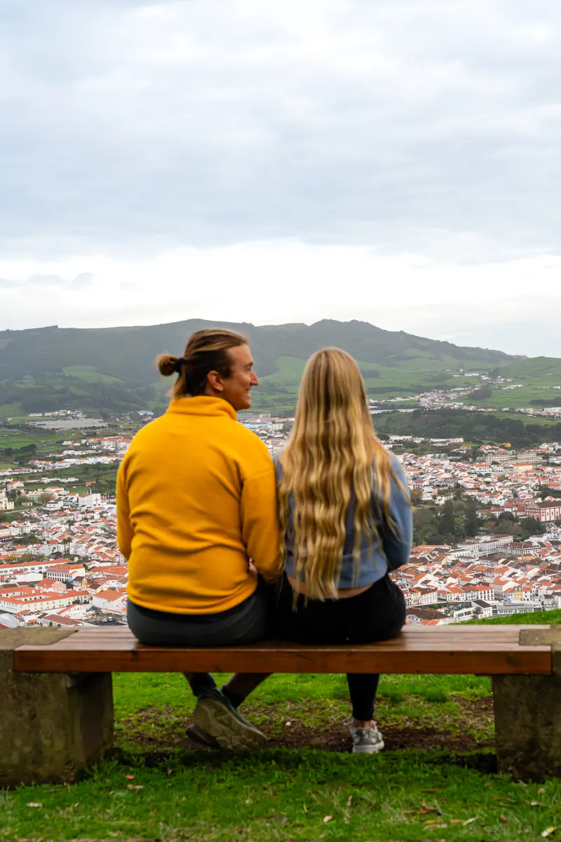 Two people sit together on a wooden bench at a hilltop viewpoint on Terceira Island, looking out over the whitewashed buildings and terracotta rooftops of Angra do Heroísmo with green volcanic hills rising behind the city.