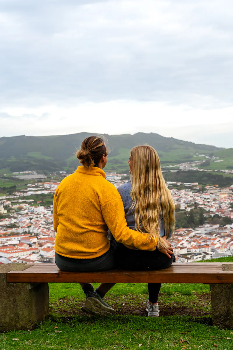 Two people sit side by side on a wooden bench at a hilltop viewpoint on Terceira Island, one with an arm around the other, looking out over the whitewashed rooftops and terracotta tiles of Angra do Heroísmo with green volcanic hills rising behind the city.