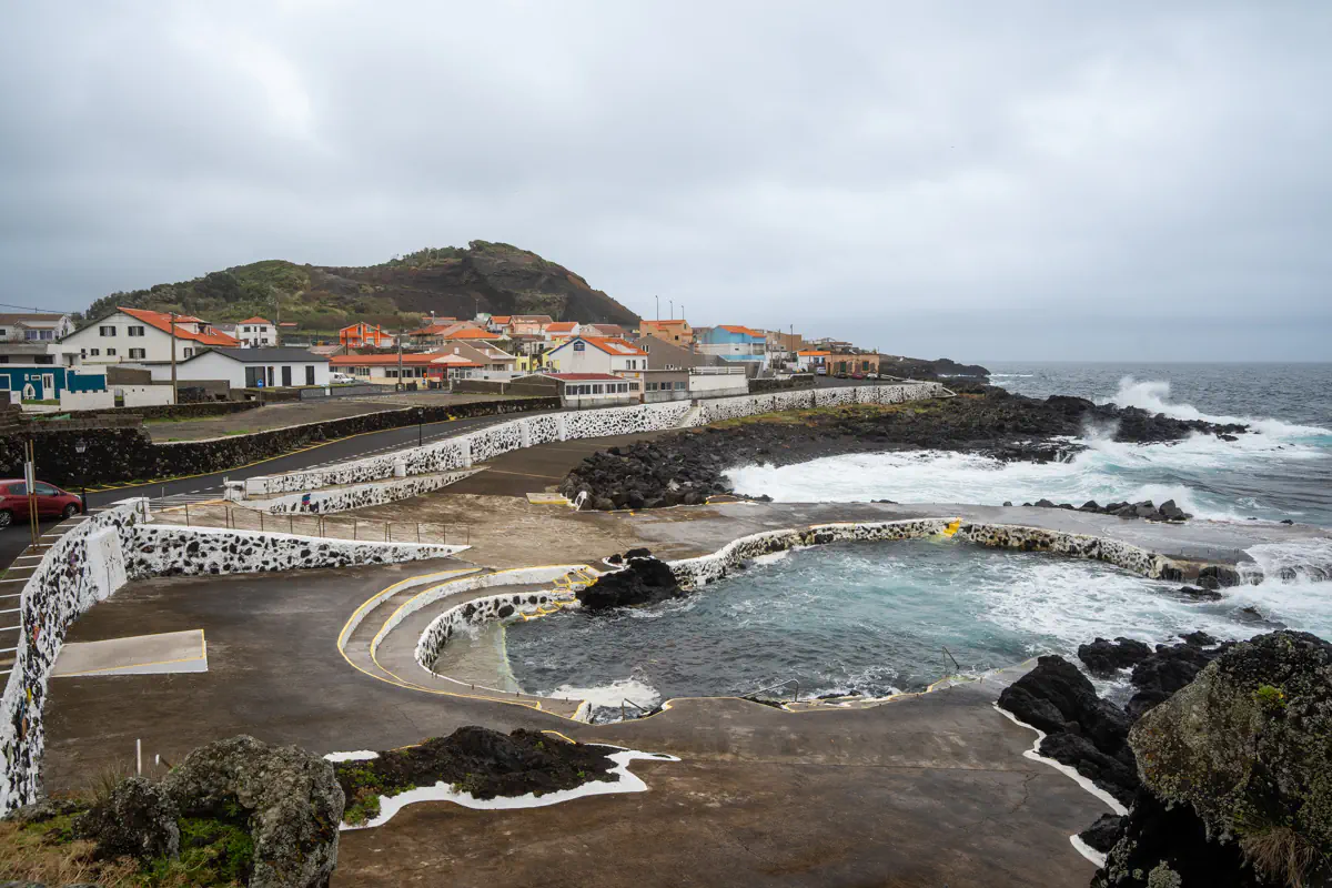 Natural lava rock pools sit beside a churning Atlantic Ocean on Terceira Island, with waves crashing over the black rocks and a small coastal village with terracotta rooftops clustered beneath a green hill in the background.